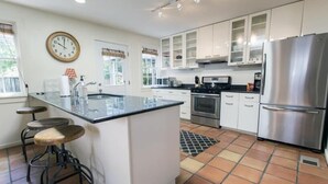 A modern kitchen with white cabinets, a stainless steel refrigerator, a central island with stools, and a clock on the wall.