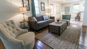 A living room with a sofa, armchair, coffee table, and a rug. There are framed pictures on the wall and a window with curtains.