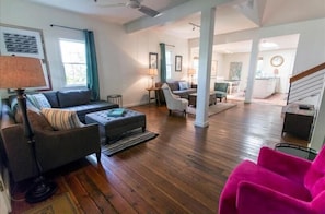 A spacious living room with hardwood flooring, a grey sofa, a bright pink chair, and a dining area in the background.