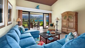A spacious living room with a large blue sofa, a wooden cabinet, and a view of a mountainous landscape through a sliding glass door.