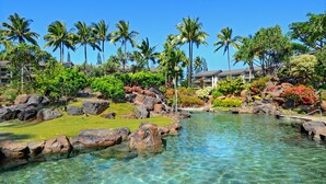 A resort with a clear pool surrounded by rocks and palm trees, and a building with multiple balconies in the background.