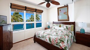 Un dormitorio con una cama grande, dos mesitas de noche, un ventilador de techo y vistas al océano a través de amplias ventanas.