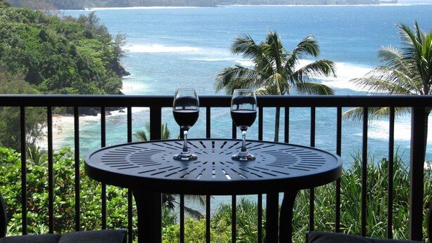 A balcony with two wine glasses, overlooking a coastal view with mountains and palm trees.