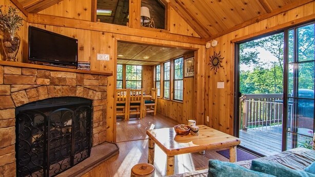 A cozy cabin interior with a stone fireplace, wooden furniture, and large windows offering a view of the outdoors. - Family Cabin on 6 Acres w/ Lake Access & Hot Tub! (Springfield)