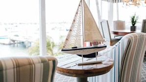 A model sailboat on a wooden table in a room with striped chairs and a view of boats through large windows.