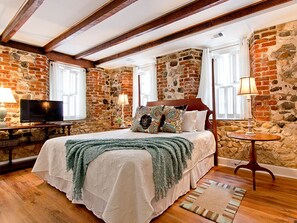 A bedroom with a stone wall, wooden beams, a bed with multiple pillows, a television, and a small round table with a lamp. - Habersham House | Historic Home, Walk To River Street (Savannah)
