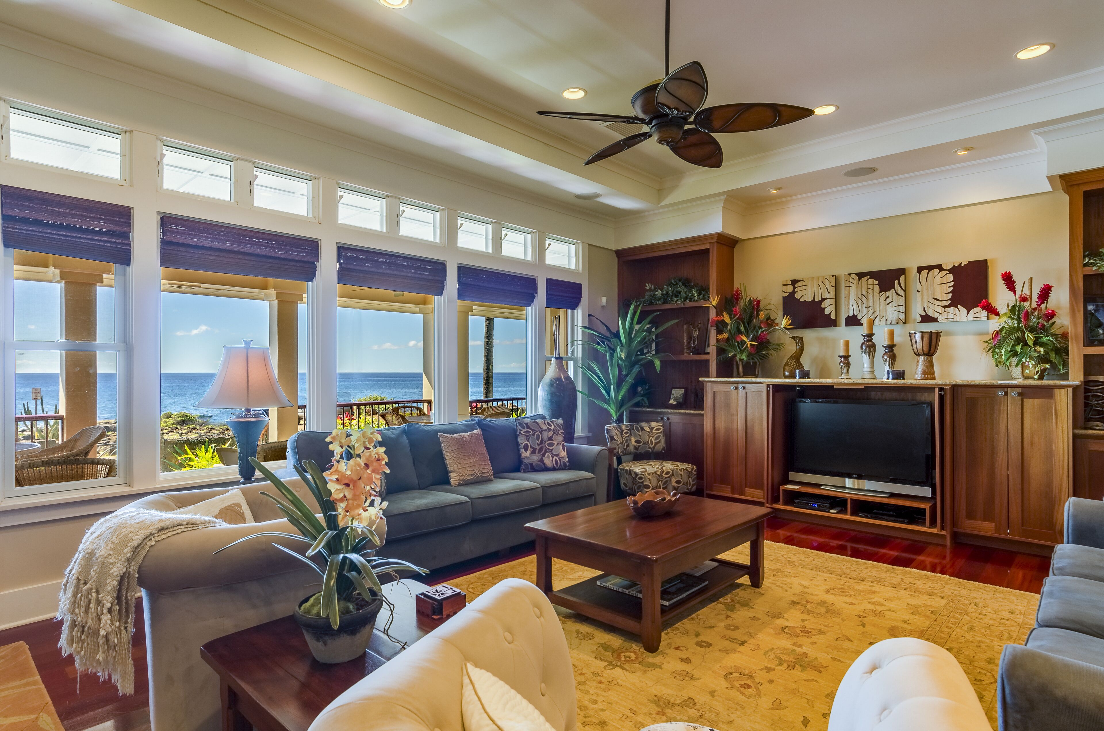 A spacious living room with a large window overlooking the ocean, a ceiling fan, and a television in a wooden entertainment unit.