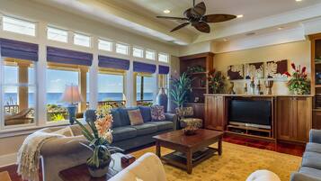 A spacious living room with a large window overlooking the ocean, a ceiling fan, and a television in a wooden entertainment unit.