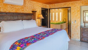 Hotelkamer met een groot bed, een kleurrijk deken, een houten hoofdbord, een wandairconditioner, een nachtkastje met een lamp, en uitzicht op een tuin door een geopende deur.