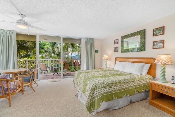 Ein Schlafzimmer mit Bett, Nachttischen, einem Sessel, einem Tisch und einem Balkon mit Blick auf Palmen.