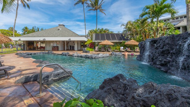 Un área de piscina con cascada, sillas de descanso y un edificio con techo de paja.