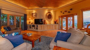 A spacious living room with a large TV, comfortable seating, a kitchen area, and a view of the outdoors.