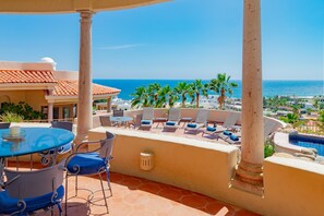 Une terrasse dotée d’une table et de chaises en verre, donnant sur une piscine et des palmiers, avec vue sur l’océan et un bâtiment dont le toit est recouvert de tuiles.