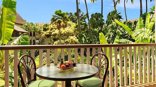 Une table ronde surmontée d’un saladier de fruits, entourée de chaises, sur un balcon doté d’une balustrade en bois.