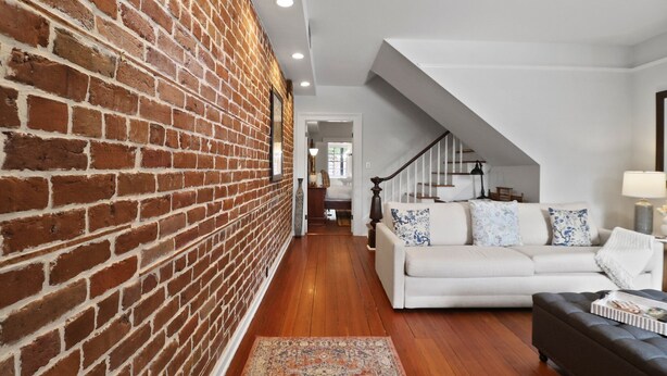 A modern living room with a brick wall, a white sofa, a dark ottoman, and a staircase leading to another level. - Garden Level Steps from Shops and Restaurants by Lucky Savannah (Savannah)