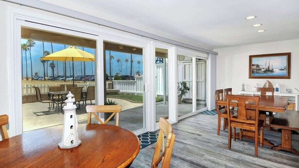 A spacious dining area with a wooden table and chairs, a yellow umbrella, and a view of the outdoor patio with palm trees and a pool. - 1005 E. Balboa A | Gorgeous Oceanfront w/ HUGE patio (Newport Beach)