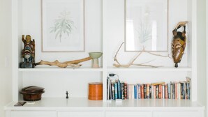 Una estantería blanca con una ilustración botánica enmarco, una escultura de madera, un cuenco de cerámica, un objeto de madera, una figurita pequeña, una colección de libros y una máscara decorativa.