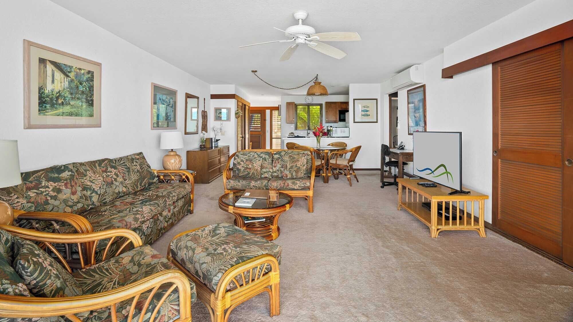 A living room with a sofa, chairs, a coffee table, and a TV. There is a dining area with a table and chairs, and a kitchen in the background.