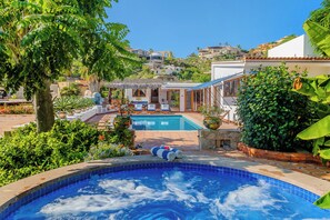 Une piscine avec un jacuzzi, entourée d’une végétation luxuriante et offrant une vue sur les bâtiments situés sur les flancs de la colline.