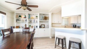A modern kitchen with white cabinets, a wooden dining table, and a ceiling fan.