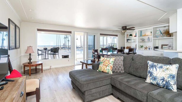 A spacious living room with a gray sectional sofa, a dining area with a table and chairs, and a view of the beach through large sliding glass doors.