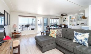 A spacious living room with a gray sectional sofa, a dining area with a table and chairs, and a view of the beach through large sliding glass doors.