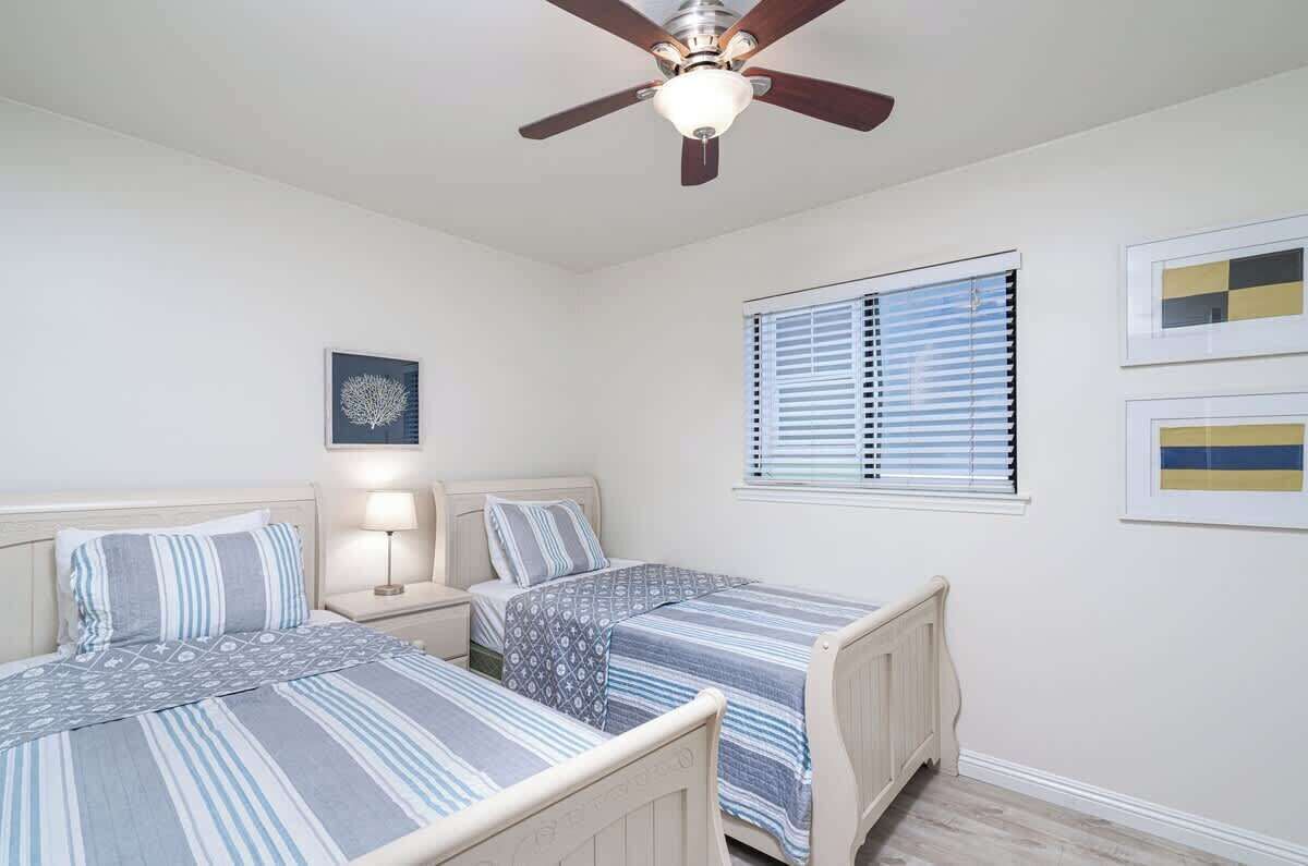 A bedroom with two beds, a ceiling fan, a nightstand with a lamp, and a window with blinds.