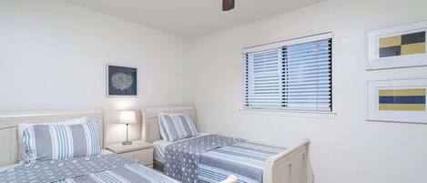 A bedroom with two beds, a ceiling fan, a nightstand with a lamp, and a window with blinds.
