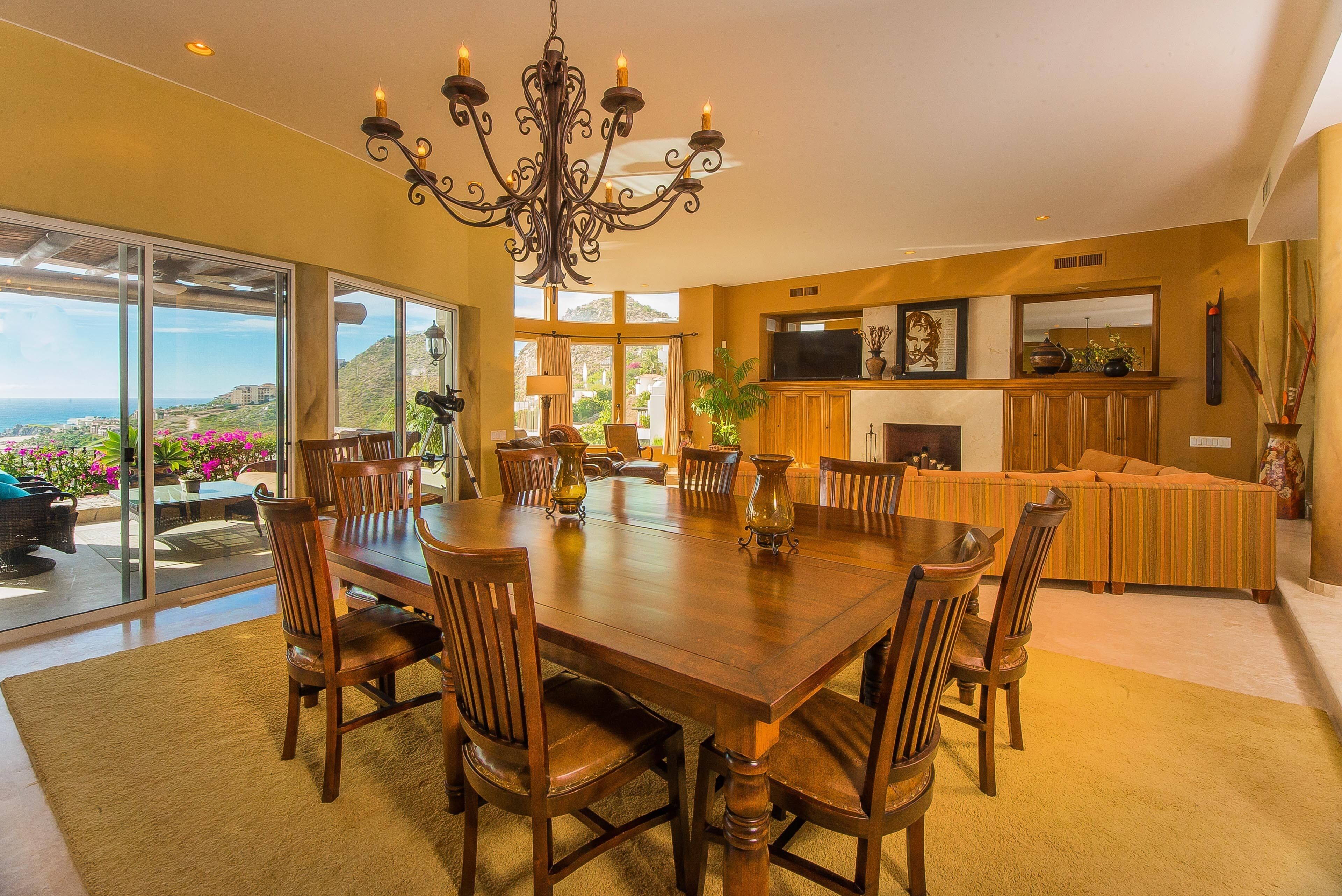 A spacious dining area with a large wooden table and chairs, a chandelier, and a view of the outdoors.