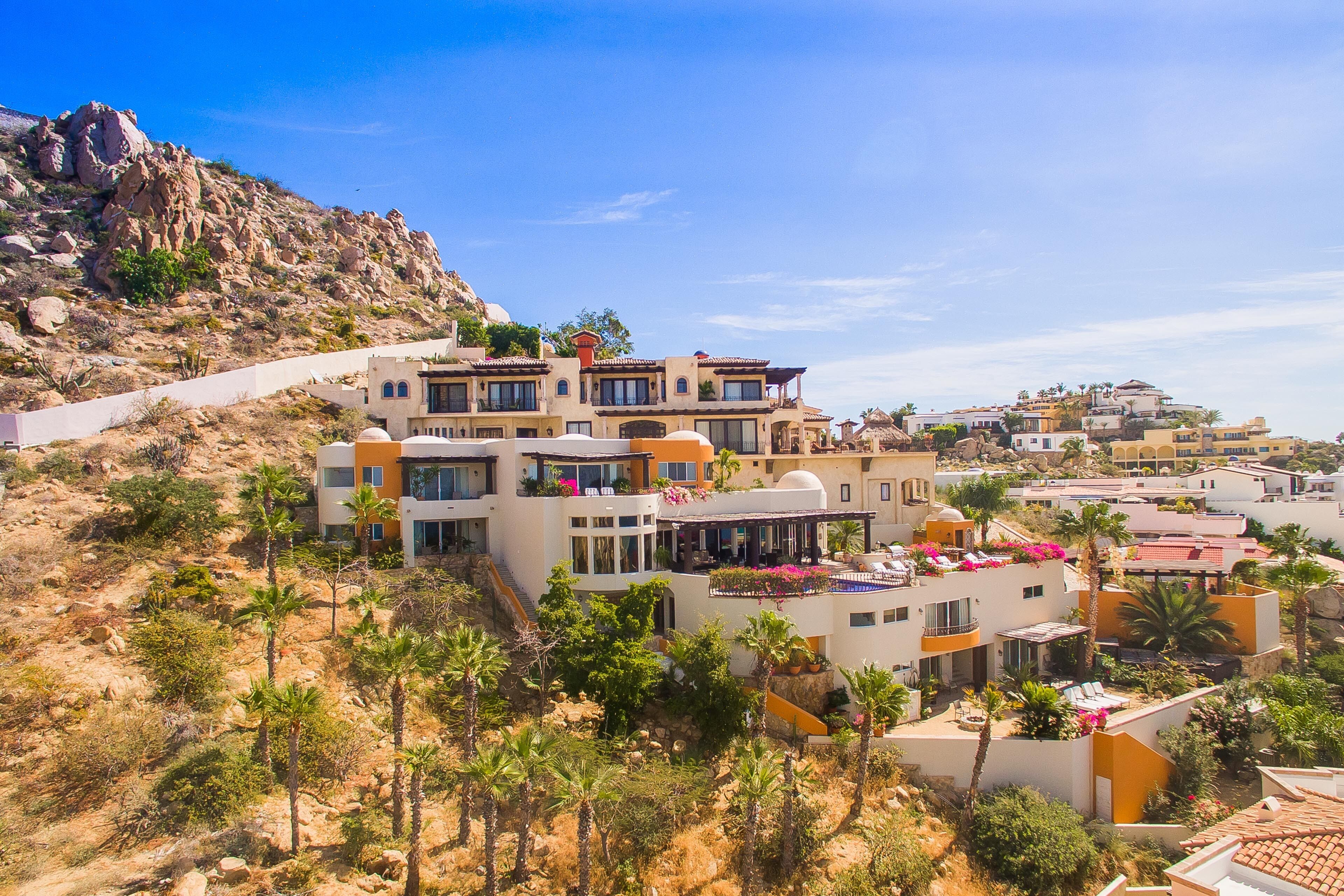A hillside residential area with modern houses featuring balconies and terraces, surrounded by palm trees and rocky terrain.