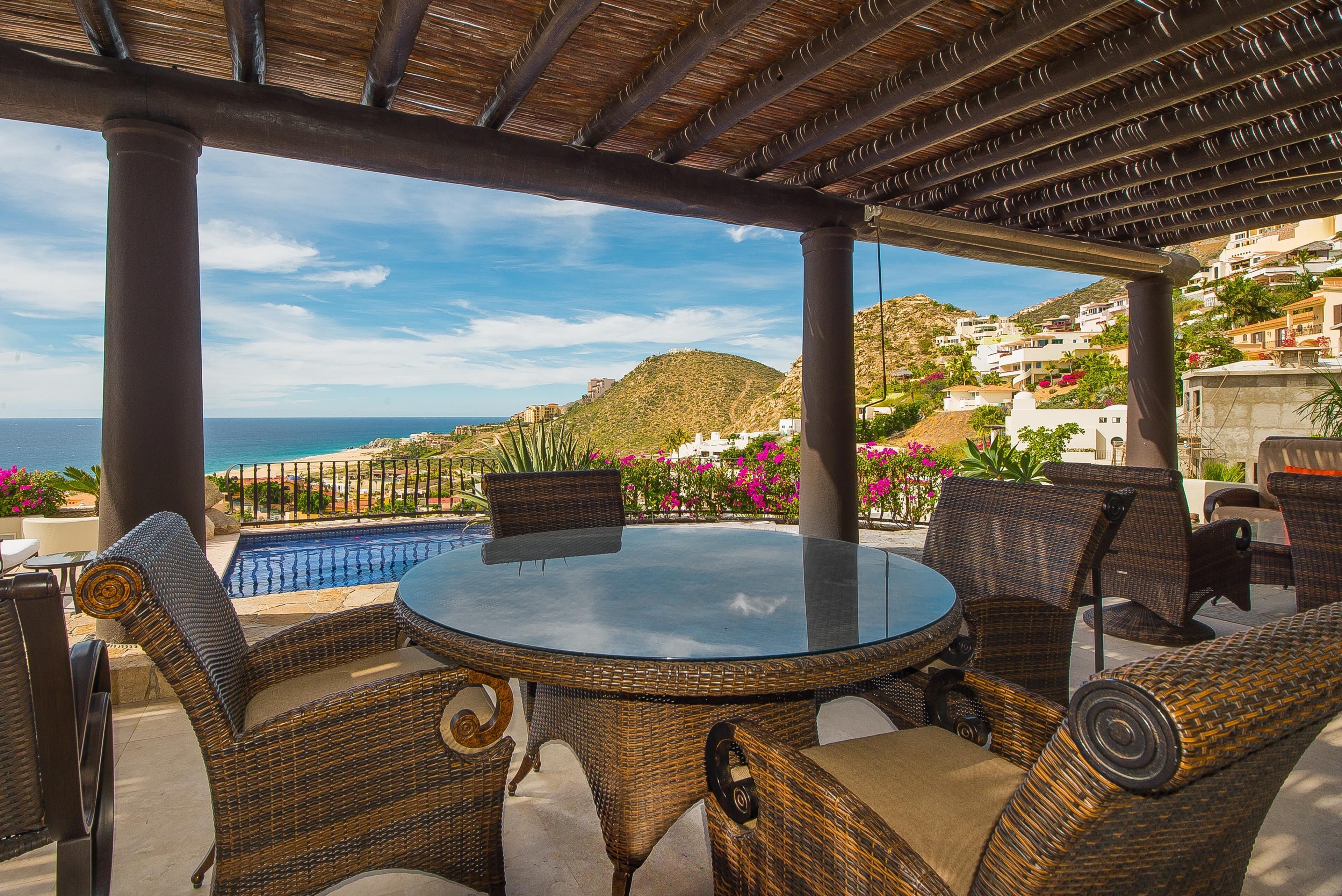 Uma área de estar coberta ao ar livre, com móveis de vime e uma mesa de vidro, com vista para uma piscina e o litoral.
