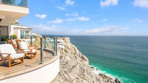 Ein Balkon am Meer mit weißen, gepolsterten Stühlen, einer Glasreling und Blick auf den Ozean.