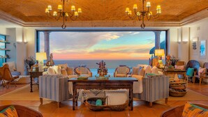 Ein geräumiges Wohnzimmer mit großem Fenster und Blick auf den Sonnenuntergang über dem Meer. Mehrere Sitzbereiche, darunter eine Sofagarnitur und Sessel, sowie ein Esstisch in der Mitte.