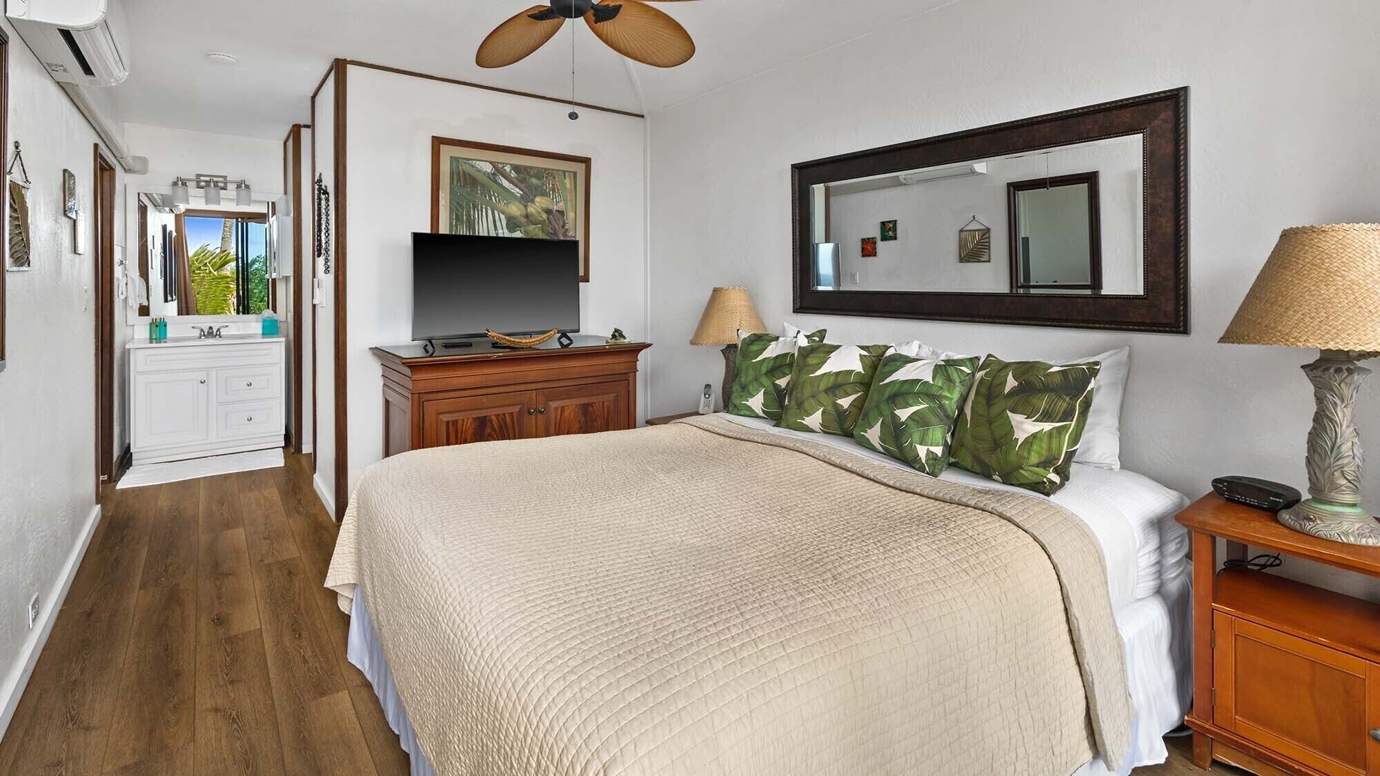 Une chambre à coucher avec un lit, une commode en bois, un téléviseur, un miroir et un ventilateur de plafond.