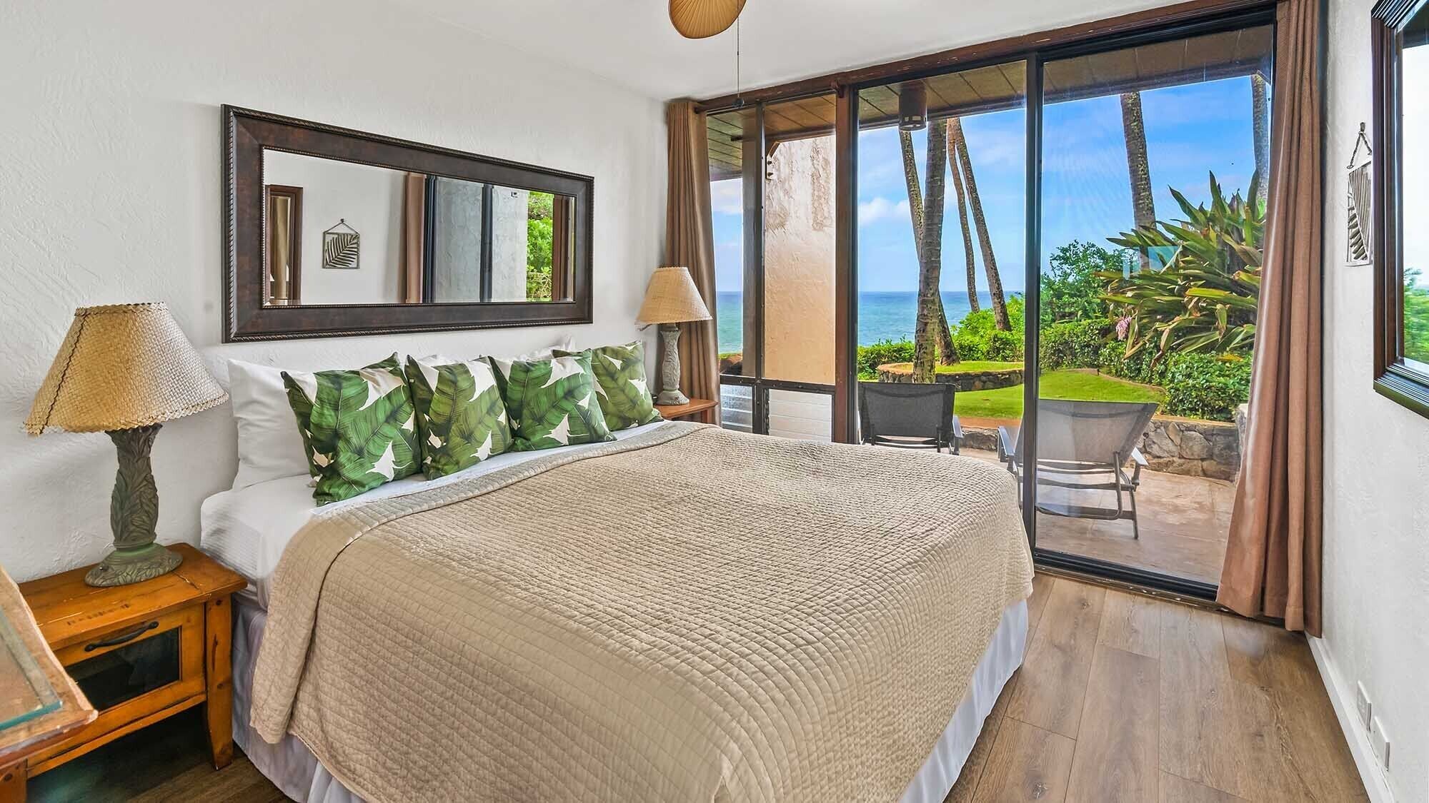 A bedroom with a large bed, a nightstand, a lamp, a mirror, and a view of the ocean through a sliding glass door.