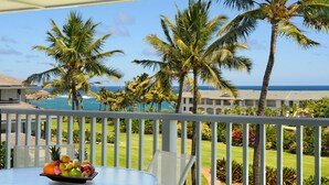 A balcony with a table holding fruit, overlooking a pool area with palm trees and a view of the ocean. - Poipu Sands at Poipu Kai #234 By Parrish Kauai - top floor ocean views & walk to (Koloa)
