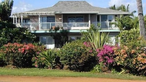 A two-story house with a balcony, surrounded by lush greenery and flowering plants. - Beach Place Duplex, Enjoy Ocean & Lush Garden Views - Poipu Beach Park (Poipu)