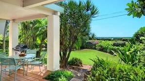 A covered patio with a grill, surrounded by lush greenery and a clear view of the ocean. - Beach Place Duplex, Enjoy Ocean & Lush Garden Views - Poipu Beach Park (Poipu)