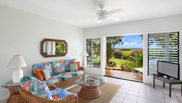 A living room with a wicker sofa, a glass coffee table, a ceiling fan, and a TV on a stand. - Beach Place Duplex, Enjoy Ocean & Lush Garden Views - Poipu Beach Park (Poipu)