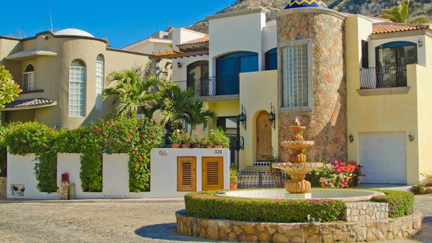Um edifício em estilo mediterrâneo, com uma fonte, paredes de pedra e montanhas ao fundo.