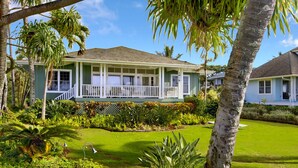 A two-story house with a porch, surrounded by lush greenery and palm trees.