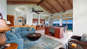 A spacious living area with a large sofa, a coffee table, and a TV. There is a kitchen area with wooden cabinets and a bar. The room has large windows with a view of the ocean.