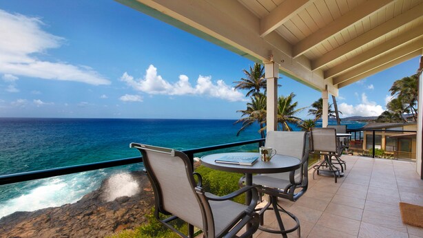 A covered outdoor seating area with a view of the ocean and palm trees.