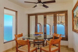 A dining area with a glass table and wooden chairs, large windows with ocean views, and a ceiling fan.
