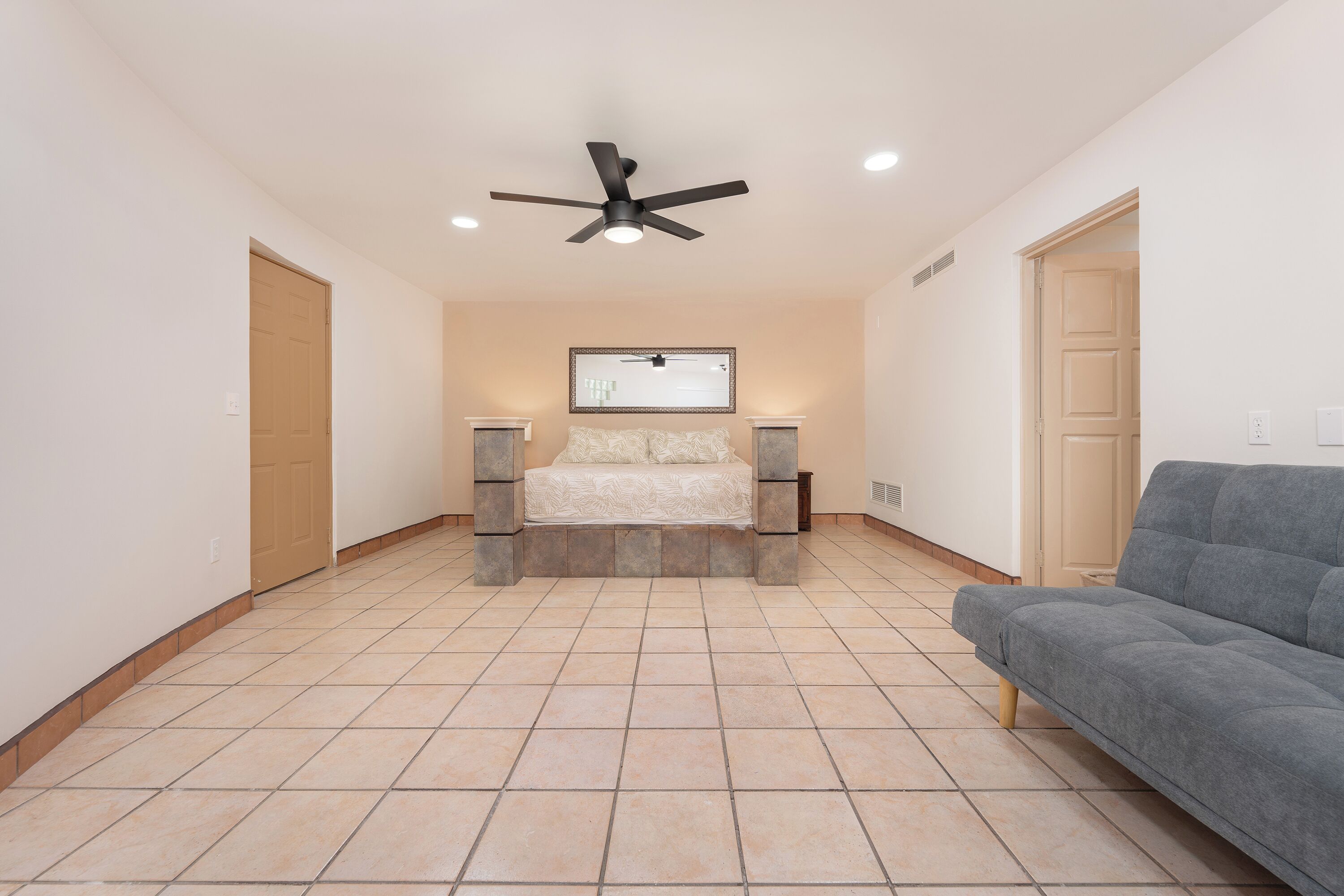 A bedroom with a bed, a ceiling fan, two bedside tables, a gray sofa, and a large mirror on the wall.