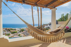 A hammock under a wooden pergola with a view of the sea and a coastal town.