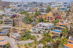 A coastal residential area with modern architecture, including houses with flat roofs and balconies, surrounded by palm trees and vegetation.