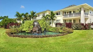 A large, two-story house with a balcony, surrounded by lush greenery and a small pond with a waterfall. - Plantation At Princeville 1521, A/C & 2nd Floor (Princeville)