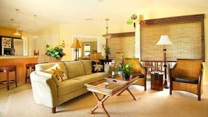 A living room with a sofa, coffee table, and wicker chairs. There is a kitchen area in the background with a bar and stools. - Plantation At Princeville 1521, A/C & 2nd Floor (Princeville)