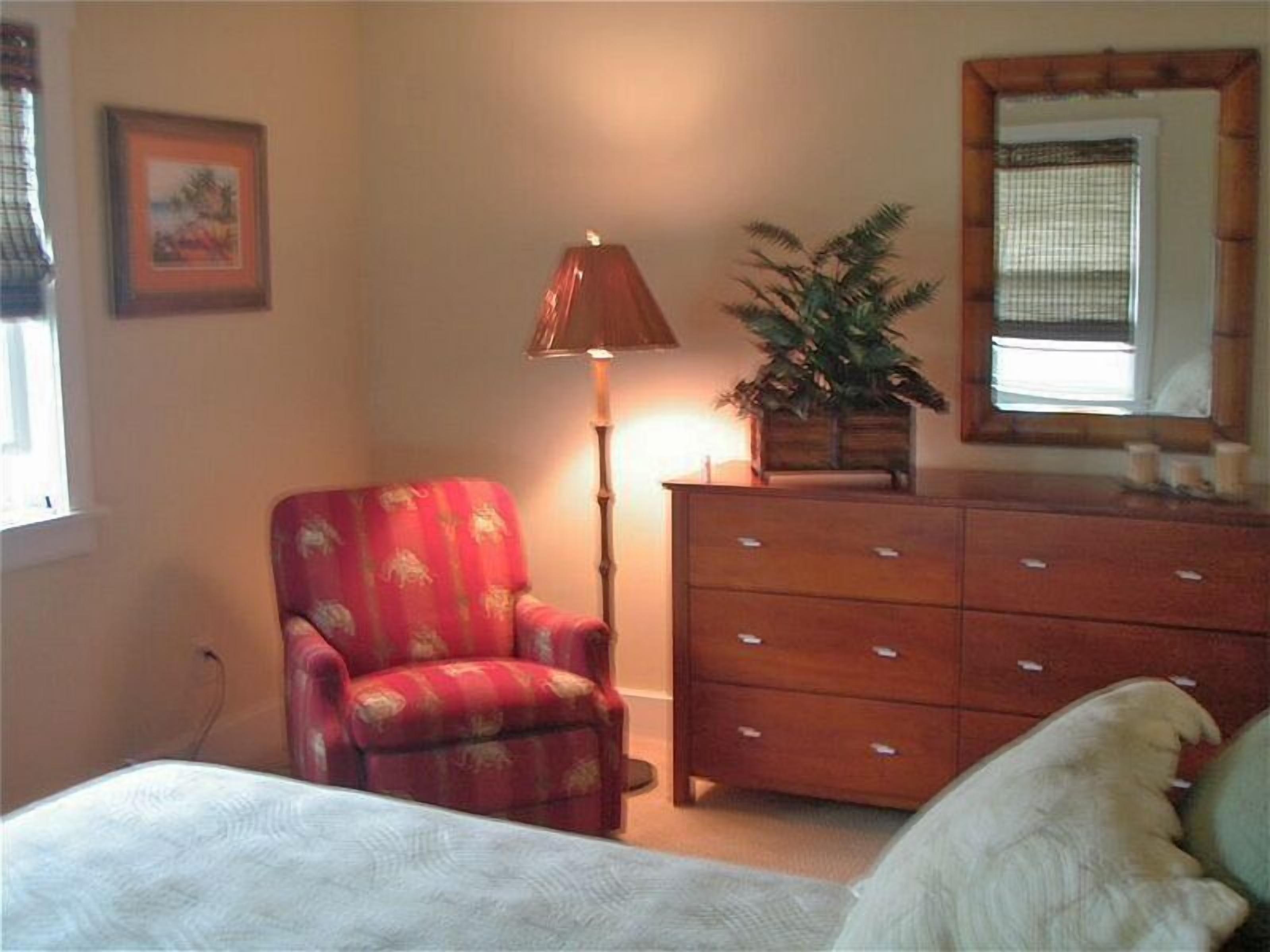 Un dormitorio con una cama, un sillón rojo, un aparador de madera, una lámpara de pie y una planta en maceta.